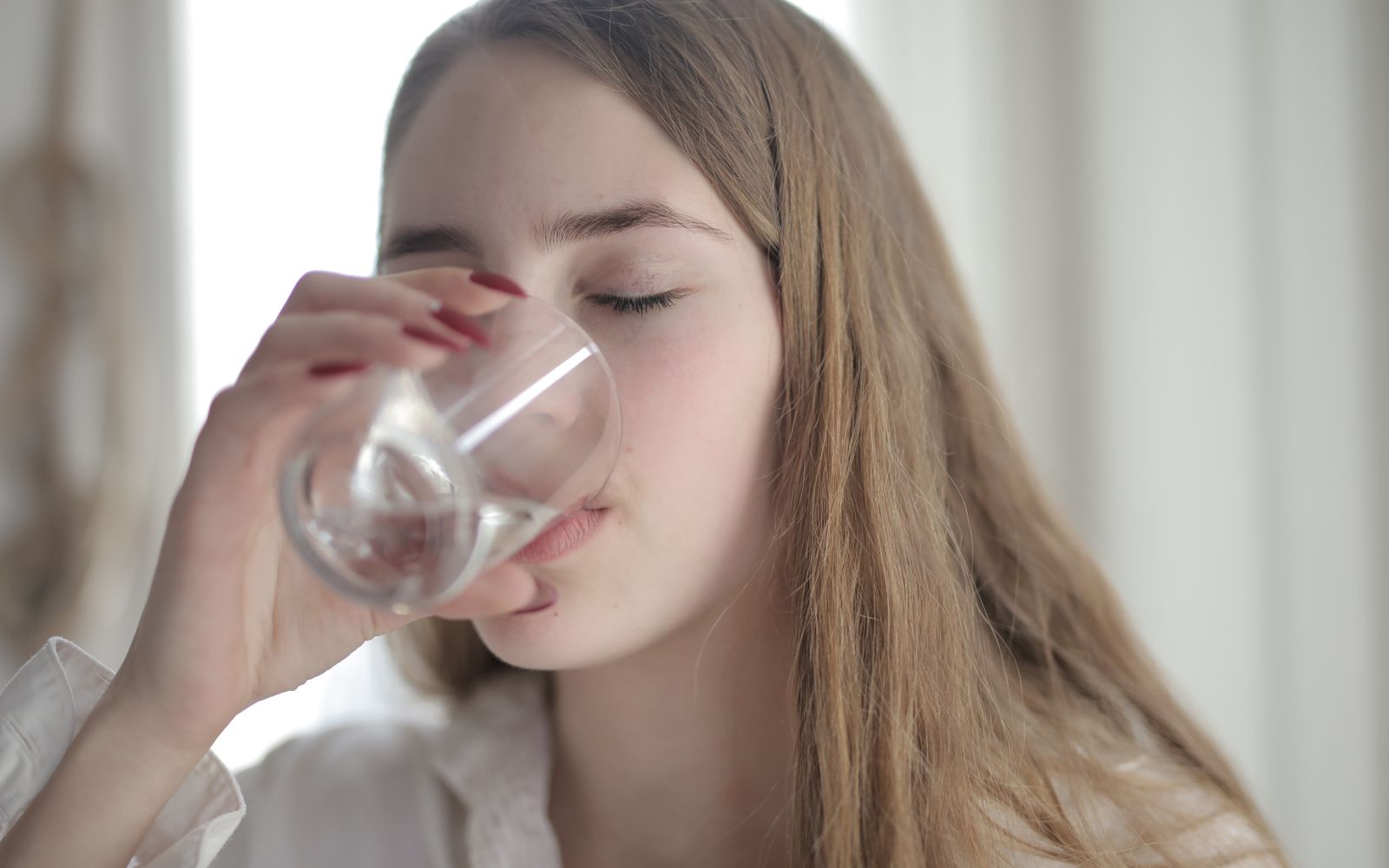 A woman in a white shirt drinking a glass of water with her eyes closed